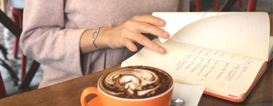 Woman Checking Journal Prompts For Goal Setting With Orange Coffee Mug